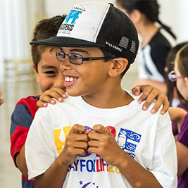 A student in a cap smiles as another student puts their hands on their shoulders from behind.