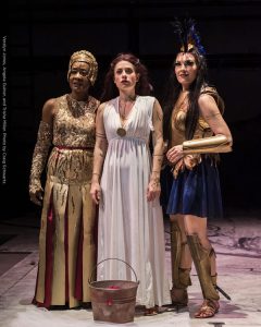 Hera, Medea, and Athena stand side by side as they look into the distance in this scene from Argonautika. Hera is wearing a gold dress, Medea is wearing a white dress, and Athena is wearing gold and dark blue warrior garb.