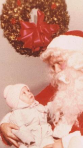 Susan Angelo as an infant in a pink jacket and beanie stares at Santa Clause with a comically disturbed look on her face as he holds her.