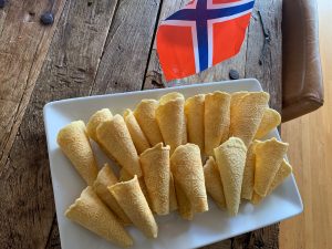 A mini Norway flag, which is red with a navy blue cross outlined with white, stands on a wood table next to a plate of beige krumkake cookies decorated with intricate hearts and rolled into cones.