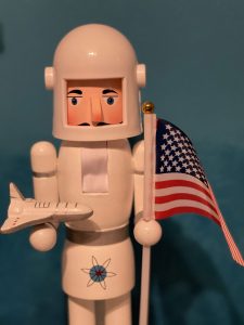 A nutcracker in a white astronaut suit stands in front of a blue background. The nutcracker holds an American flag in one hand and on the other hand holds a narrow white and silver space ship with three fins.