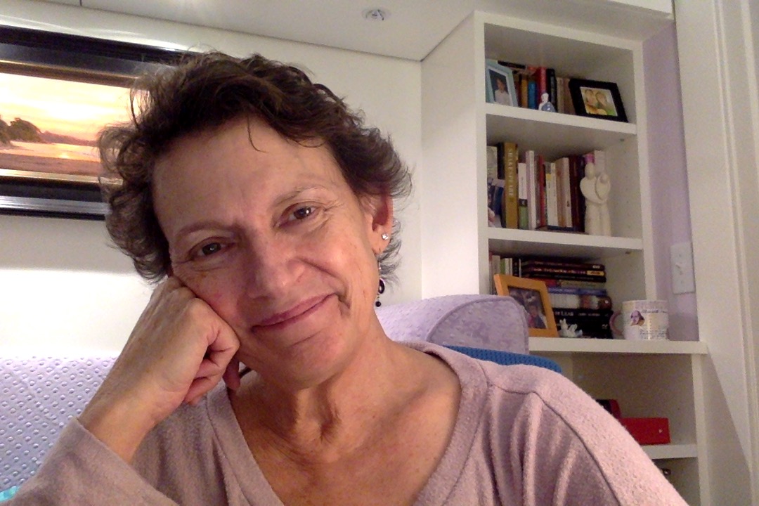 Miranda has short curly brown hair and wears a mauve shirt as she leans her head against her hand and smiles. She is sitting in a brightly lit white room with a bookshelf behind her.
