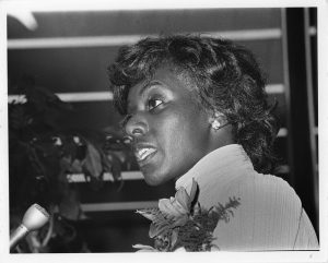 A black and white photo of Loretta Thompson-Glickman giving a speech in front of a mic. She is a Black woman with short wavy dark hair and wearing a high-collared striped shirt.