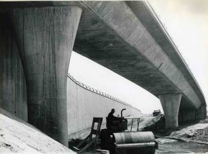 A black and white photo of a construction worker in a hard hat operating a steam roller under a bridge of the 210 freeway.