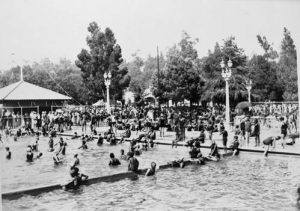 A black and white photo of the Brookside Plunge pool with large crowds of people swimming and playing and walking around. Several lampposts pepper the backdrop and large trees dominate the background landscape.