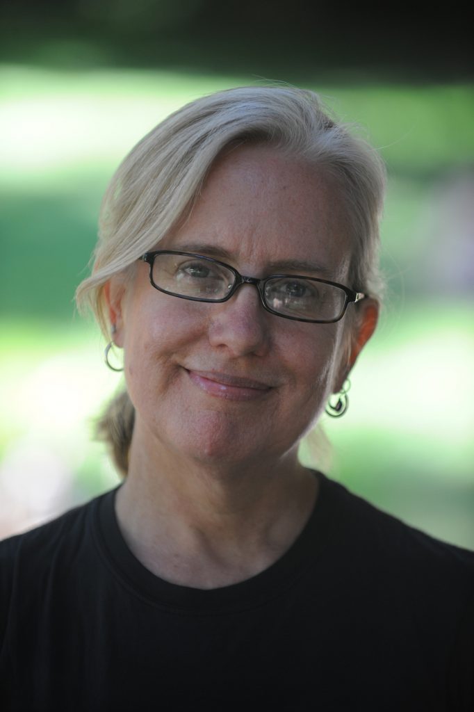 Lisa Peterson has blond hair tied back and black glasses and a black T shirt. She smiles in front of a blurred green background.