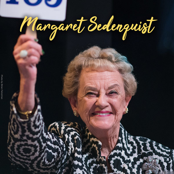 Margaret Sedenquest in yellow cursive. Margaret Sedenquist pictured at Dinner On Stage as she smiles and holds up a paddle. She has short curly blond hair with a green streak and wears a coat with black and white flowers.