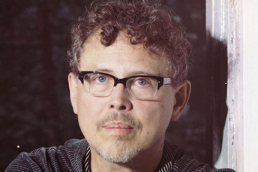 Jose Rivera has brown curly hair and faint mustache and beard and wears black rimmed glasses.